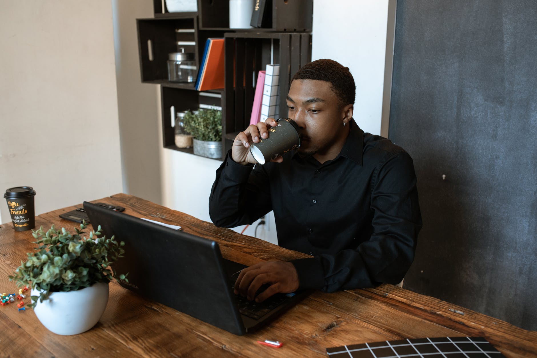 man in black long sleeves drinking cup of coffee while busy looking at the screen of laptop